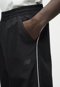 Άτομο με μαύρο αθλητικό σορτς με ελαστική μέση και λευκή πλευρική γραμμή, το χέρι μέσα στην δεξιά τσέπη, ορατό το λογότυπο New Balance.