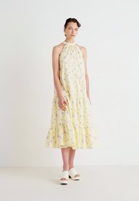 Žena v bezrukávových žltých kvetinových šatách s halter krkom a bielych vysokých platformových sandáloch stojí na plain bielom pozadí.