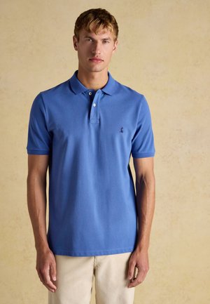 Young man wearing a blue short-sleeve polo shirt and light beige pants standing against a plain beige background.