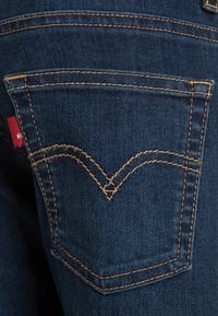 Los jeans de mezclilla de color azul oscuro presentan un bolsillo trasero con costuras doradas y una etiqueta con el logo en rojo. El bolsillo tiene un diseño distintivo en forma de V.