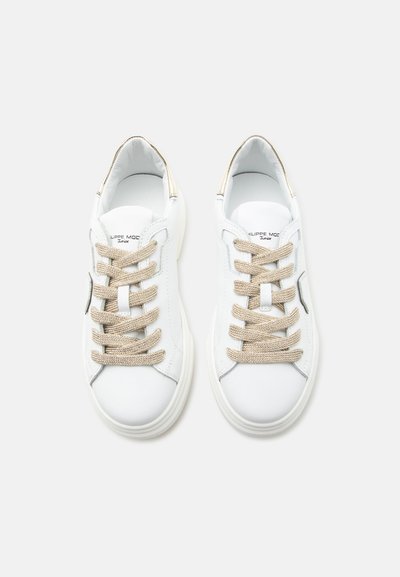 Paire de baskets basses blanches avec lacets beige, texte de marque visible à l'intérieur et subtils accents métalliques sur talons et côtés.