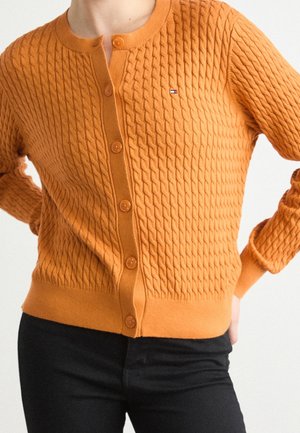 Frau trägt eine strukturierte orangefarbene Strickjacke mit Zopfmuster und Knöpfen, mit einem kleinen Flaggenlogo auf der Brust, kombiniert mit schwarzen Hosen.