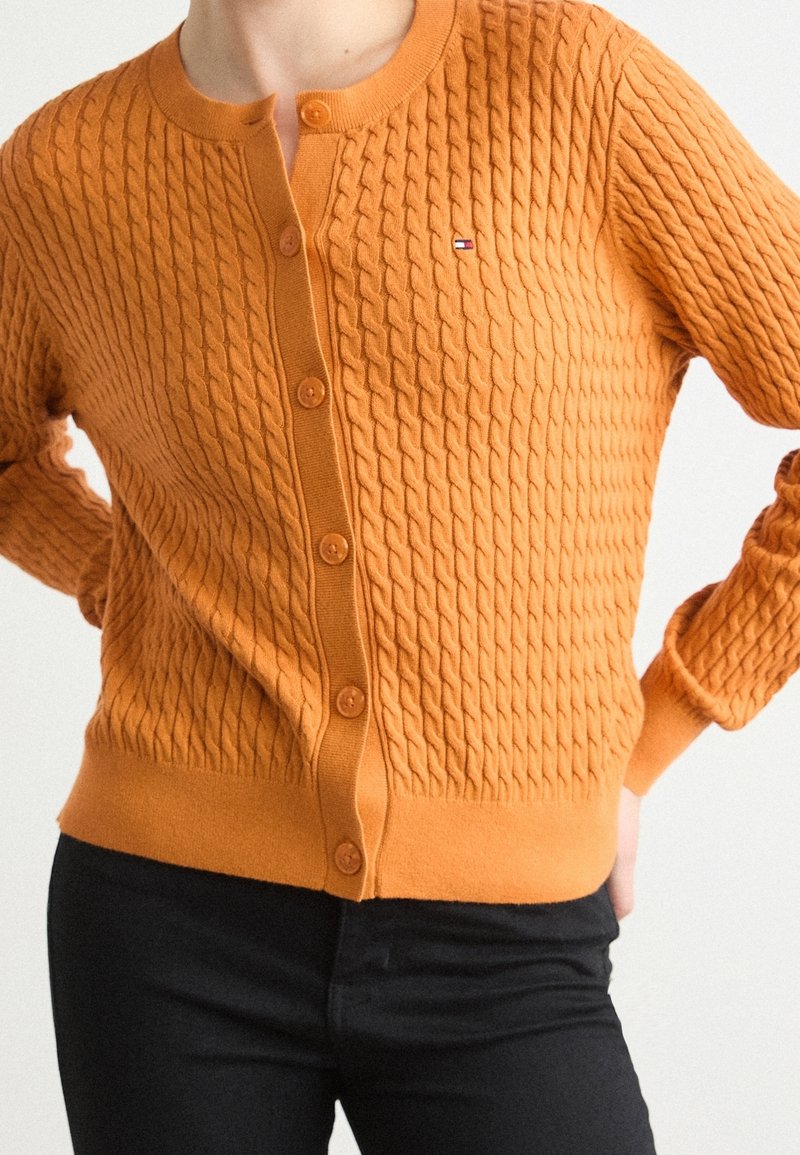 Femme portant un cardigan en maille torsadée orange texturée à boutons avec un petit logo de drapeau sur la poitrine, associé à un pantalon noir.