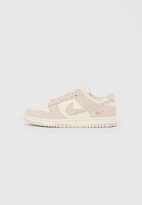 Nízký Nike teniska v béžové a krémové barvě s semišovými panely, tkaničkami a malým vyšívaným oranžovým znakem Swoosh na patě.