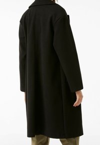 Černý vlněný kabát s vysokým límcem, dlouhými rukávy a rovnou siluetou. Má boční kapsy a délku po lýtka. Texturovaná látka.