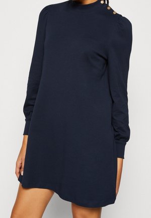 Kvinde iført en mørkeblå kjole med lange ærmer, guldknapper på skulderen og høj hals, stående mod en ensfarvet baggrund.