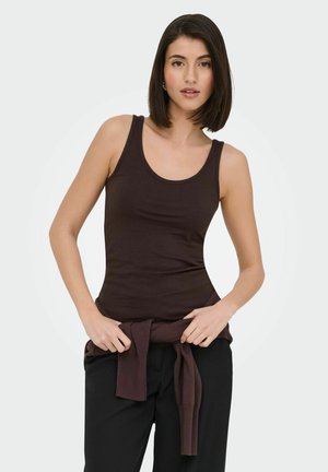 Young woman with shoulder-length dark hair wearing a dark brown tank top and tying a matching sweater around her waist.
