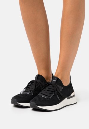 Czarne dzianinowe trampki z grubymi białymi podeszwami noszone na bosej stopie, widoczne z przodu i z boku na jasnym, jednolitym tle.