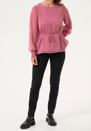 Femme portant un chemisier rose transparent à manches longues avec un lien à la taille, pantalon noir ajusté et mocassins noirs, debout devant un fond blanc.