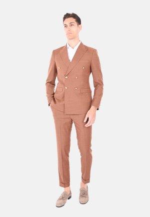 Completo marrone doppiopetto in tessuto testurizzato, con due tasche frontali e bottoni dorati. Abbinato a una camicia bianca e mocassini in suede chiaro.