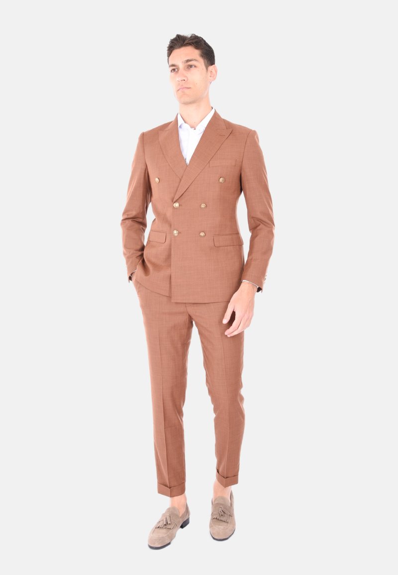Completo marrone doppiopetto in tessuto testurizzato, con due tasche frontali e bottoni dorati. Abbinato a una camicia bianca e mocassini in suede chiaro.
