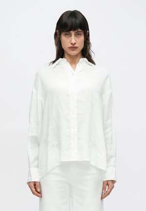 Biela dlhá košeľa z ľanového materiálu s gombíkmi na prednej strane, špicatým golierom a voľným strihom. Lem je mierne zaoblený a rukávy sú vyhrnuté.