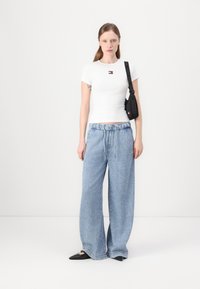 Hvit ribbet t-skjorte med kort erm og brodert logo, kombinert med lyseblå vide jeans og svarte spisse leiligheter, holder en liten svart veske.