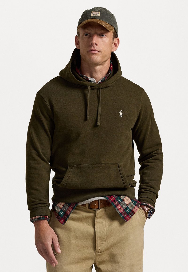 Sweat à capuche vert olive avec cordons et poche avant, superposé sur une chemise à carreaux rouge et vert, associé à un pantalon beige et une ceinture marron.