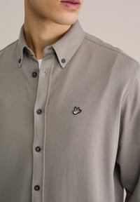Camisa abotonada en un tejido texturizado gris claro con un sutil patrón de rayas, con botones negros y un logo bordado en el pecho.