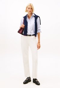 Frau in weißen Hosen, gestreiftem hellblauen Hemd, mit einem marineblauen Pullover über den Schultern, schwarzem Gürtel, schwarzen Slippern und einer weinroten Handtasche.