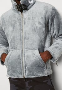 Szara kurtka polarowa z wysokim kołnierzem, przednim zamkiem błyskawicznym i podwójnymi klamrami dekoracyjnymi. Długie rękawy oraz boczne kieszenie zwiększają funkcjonalność. Miękka w dotyku.