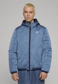 Blå polstret jakke med hette, med glidelås foran og logo på brystet. Jevn tekstur, elastiske mansjetter og kant.