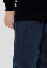Zwarte sweatshirt met geribbelde manchetten, voorzien van witte tekst op de mouw. Een paar donkerblauwe denim jeans met een gladde textuur.