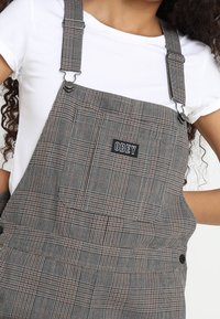 Graue karierte Latzhosen mit schwarzen Trägern, die eine Fronttasche mit der Aufschrift "OBEY" und seitlichen Knöpfen zur Anpassung aufweisen.