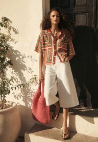 Camisa abotonada multicolor de crochet con patrones geométricos, combinada con pantalones blancos de pierna ancha y un bolso tote tejido rojo. Chanclas negras.