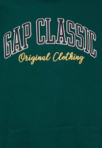 Felpa verde con cappuccio caratterizzata dalla scritta "GAP CLASSIC" in lettere bianche in grassetto ricamate, con "Original Clothing" in corsivo dorato sotto.