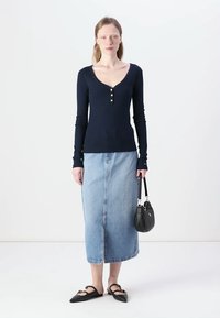 Top cu mânecă lungă de culoare bleumarin, cu trei nasturi aurii la gât, asortat cu o fustă din denim albastru deschis și balerini negri, ținând o mică geantă neagră.
