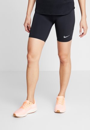 Persoon draagt zwarte Nike-fietsshorts en lichtroze sportschoenen, staand op een grijze vloer.