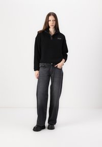 Zwarte fleece pullover met een kraag, voorzien van een logo op de borst, gecombineerd met losse grijze jeans en zwarte chunky laarzen.