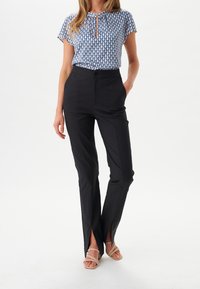 Femme portant un haut à manches courtes à motifs bleus, un pantalon noir taille haute avec des fentes à l'avant, et des sandales à lanières beiges, se tenant avec une main dans la poche.