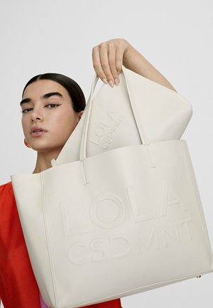 Žena s tmavými vlasmi oblečená v červenej farbe drží veľkú béžovú tašku a zodpovedajúci púzdro s embosovaným nápisom "LOLA CSDMNT" na bielom pozadí.