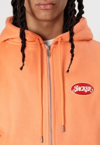 Orange zip-up hoodie made of soft fabric, featuring a logo patch, silver zipper, and drawstrings with metal tips.