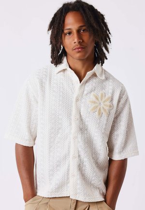 Homme avec dreadlocks portant une chemise blanche texturée à manches courtes avec un grand motif de fleur beige sur la poitrine gauche.
