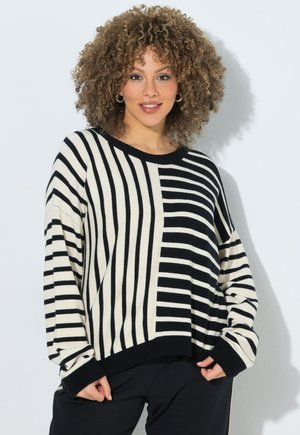 Kvinde med krøllet hår iført sort og hvid stribet sweater står mod en hvid væg, smilende og holder i sweaterens kant.