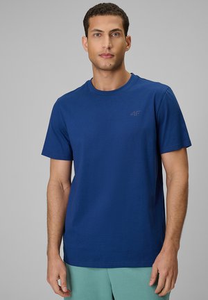 Mężczyzna w gładkim niebieskim t-shircie z krótkim rękawem i jasnozielonych spodniach stoi na tle szarego tła.