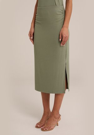 Femme portant une jupe midi côtelée vert olive avec une fente sur le côté et des sandales à talons hauts à brides couleur beige.