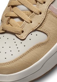 Béžové tenisky s texturovaným syntetickým koženým svrškem, bílou špičkou a kulatými perforacemi. Obsahují béžové tkaničky a polstrovaný jazyk pro pohodlí.