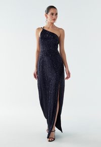 Námornícky modré večerné šaty so sieťovaným zdobením, s jedným ramenným popruhom, vysokým rozparkom až po stehná a hladkou textúrou, doplnené čiernymi páskovými sandálmi.