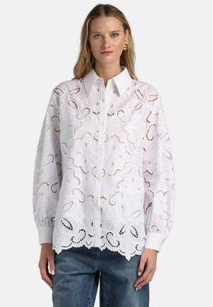 Kobieta ubrana w białą, kwiatową bluzkę z ażurowej koronki z długimi bufkami, zapinaną na guziki z falbanką na dole, w zestawieniu z niebieskimi dżinsami.