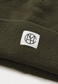 Olijfgroene gebreide beanie met een omgeslagen zoom en een wit label met een zwart logo. Zachte textuur, dichtgebreid ontwerp.