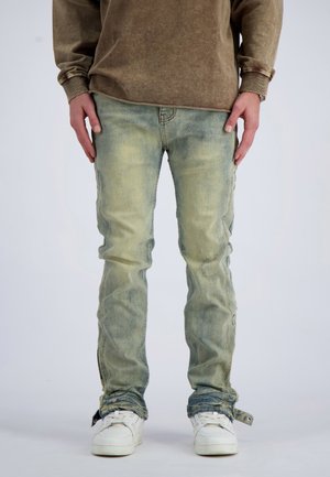 Person trägt verblasste blaue Jeans mit Reißverschlüssen an den Knöcheln, weiße Turnschuhe und ein braunes Langarmshirt und steht vor einem schlichten Hintergrund.