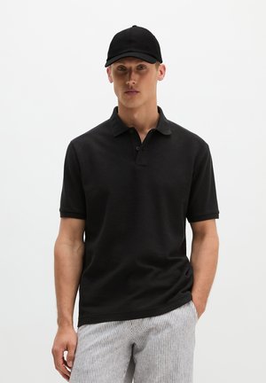 Man draagt een zwart poloshirt en een zwarte pet, met lichtgekleurde gestreepte broek, staand tegen een effen witte achtergrond.