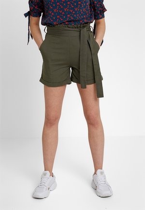 Person iført olivengrønne, højtaljede shorts med bundet bælte, en marineblå blomstermønstret bluse og hvide sneakers, stående med hænderne i lommerne.