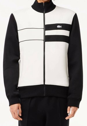 Mann trägt eine schwarz-weiße Jacke mit Reißverschluss, schwarzen Ärmeln und zwei horizontalen schwarzen Streifen auf der Brust, mit einem kleinen Krokodil-Logo.