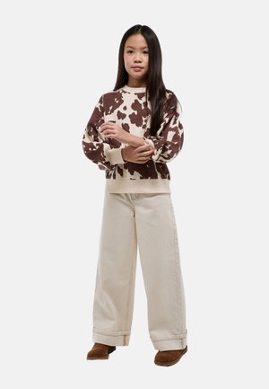 Jeune fille aux longs cheveux portant un pull marron et crème à motif vache, un pantalon large crème et des chaussures marron, debout les mains jointes.