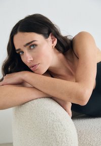Model with long dark hair, wearing a black garment, resting on a textured light-colored, rounded surface. Focus on facial features and skin tone.