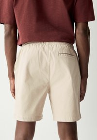 Shorts beiges avec une ceinture élastique, des poches latérales et une poche arrière avec un bord contrastant. Le tissu a une texture lisse.