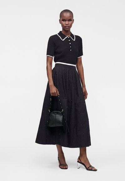 Femme portant un haut noir à manches courtes avec col et une longue jupe noire ornée, tenant un sac à main noir et portant des talons noirs à lanières.