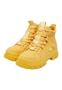 Zapatillas altas amarillas de tela con un acabado texturizado, que cuentan con suelas de goma gruesas, cordones y una marca en la lengüeta y el talón.