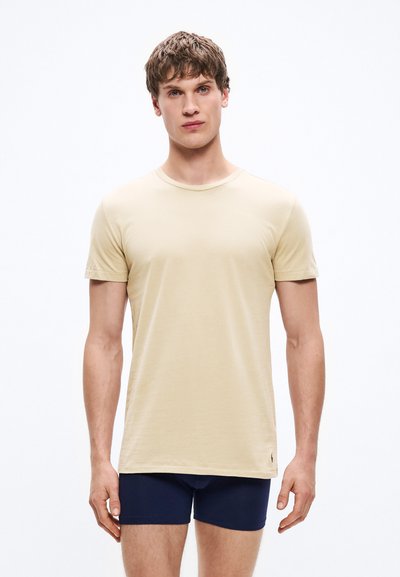 Μπεζ βαμβακερό t-shirt με κοντά μανίκια, στρογγυλή λαιμόκοψη και διακριτική λεπτομέρεια λογότυπου στο τελείωμα. Το μοντέλο φοράει ναυτικές βερμούδες.